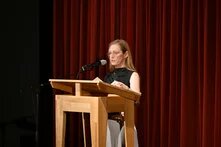 Therese Berg steht an einem Holzpult und spricht in ein Mikrofon. Im Hintergrund hängt ein roter Vorhang.