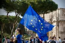 Rome, Italy. A person is waving a large EU flag in front of the Arch of Constantine. Many people are standing around them, some in blue clothing.