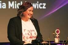 Foto: Eine lachende Frau steht an einem Rednerpult. Im Hintergrund ist ein Bildschirm mit Text, daneben der Anne-Klein-Frauenpreis