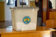 Eine transparente Wahlurne mit dem Siegel der Zentralen Wahlkommission Moldaus steht auf einem Holztisch. Sie enthält ausgefüllte Stimmzettel. Im Hintergrund sind eine Tür und eine Person sichtbar.