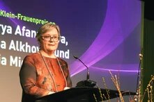 Photo: A woman with short gray hair stands at a lectern. A screen with text and a purple graphic design is in the background.