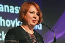 Photo: A woman with short red hair speaks into a microphone. A screen with text and purple graphic design is in the background.