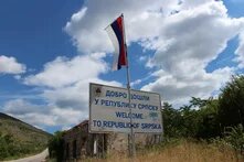 Foto: Grenzschild zur Republika Srpska mit serbischer Flagge, davor eine Straße, dahinter ein altes Steingebäude und bewachsene Hügel.