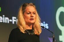 Photo: A woman with long blonde hair speaks into a microphone. A screen with partially readable text is in the background.