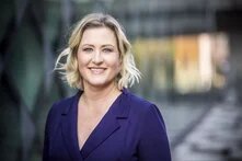 Eine Person mit blondem, schulterlangem Haar steht draußen und lächelt in die Kamera, im Hintergrund Glasfassade und Weg.