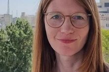 Foto: Eine Person mit langen, glatten Haaren und Brille steht im Freien vor Bäumen und Gebäuden im Hintergrund.