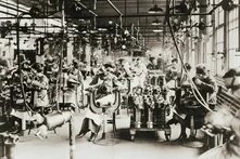 Foto: Innenaufnahme einer historischen Fabrikhalle mit vielen arbeitenden Personen an Maschinen in eng gestellten Reihen