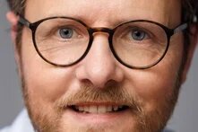 Foto: Porträt einer Person mit Brille und Bart vor neutralem Hintergrund, Blick direkt in die Kamera. 