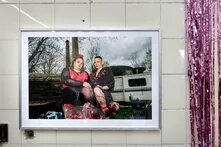 Foto: Gerahmtes Porträt zweier Personen vor Wohnwagen und Bäumen, ausgestellt an gefliester Wand mit pinkem Lamettavorhang 
