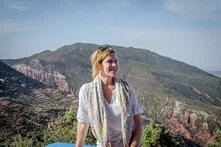 Antonie Nord steht vor bergiger Landschaft mit grünen Hängen und rotbraunen Felsen, sie trägt ein helles Oberteil und schaut lächelnd leicht zur Seite.