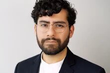 Person with glasses, beard, and curly hair looks seriously at the camera against a light background.