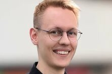 Eine lächelnde Person mit Brille blickt in die Kamera, kurzer Haarschnitt, unscharfer heller Hintergrund.