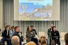 Mehrere Personen sitzen in einem Raum mit Stuhlkreis. Eine Person spricht ins Mikrofon. Im Hintergrund zeigt eine Leinwand Bilder mit dem Titel „Jasenovac and Bleiburg“.