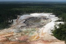 Große offene Mine mit mehreren Baggern am Rand eines dichten Waldgebiets, Boden zeigt verschiedene Farbnuancen.
