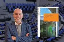 Eine lächelnde Person in Anzug und mit Brille steht mit verschränkten Armen vor einem leeren Plenarsaal; rechts ein Buchcover mit Titel "Strukturen des Fortschritts".
