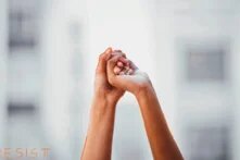 Two raised hands clasp each other, blurred buildings in the background; ‘RESIST’ is written at the bottom.