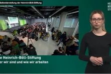Großer Sicht in einen Saal mit Publikum, Gebärdensprachdolmetscherin rechts im Bild.
