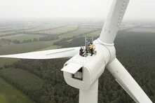 Mehrere Personen stehen gesichert auf dem Maschinenhaus einer Windkraftanlage über bewaldeter Landschaft.