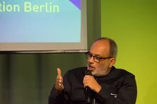 A man with glasses speaks into a microphone and makes gestures with his left hand; behind him is part of a screen with the word “Berlin”.