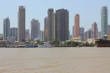 Several high-rise buildings behind the riverbank with a boat and houses in the foreground, smoke rising between the trees.