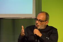 A man with glasses speaks into a microphone and makes gestures with his left hand; behind him is part of a screen.