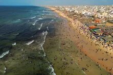 Menschenmengen im Wasser und am breiten Strand, dicht gedrängte Gebäude rechts, kleine Wellen links im Meer.