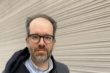 Person mit Brille und Bart steht vor einer Wand mit waagerechten Rillen, Blick zur Kamera.