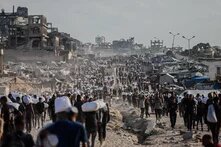Tausende Mensch*en mit Säcken auf staubiger Straße zwischen Zelten und zerstörten Gebäuden.