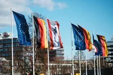 Europa-, Deutschland- und Berlin-Flaggen an Fahnenmasten wehen vor Gebäuden im Hintergrund-