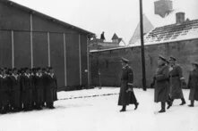 Gruppe uniformierter Frauen steht in Reihen, mehrere Männer in Uniform gehen vorbei, Schneeboden, hoher Zaun mit Stacheldraht im Hintergrund.
