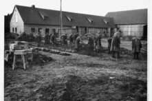 Gruppe gestreift gekleideter Personen gräbt mit Schaufeln im schlammigen Hof vor langem Backsteingebäude mit Scheunen.