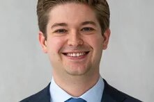 Ein junger Mann mit kurzen, gestylten Haaren lächelt und blickt frontal in die Kamera. Er trägt Anzug, Hemd und Krawatte.