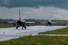 Zwei Kampfjets rollen hintereinander auf nasser Landebahn, Wiese und Wald rechts und links, dunkle Wolken am Himmel.