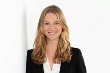 Eine Person mit langen, welligen Haaren steht vor hellem Hintergrund und lächelt in die Kamera.