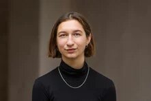 Person mit kinnlangem Haar blickt frontal in die Kamera, im Hintergrund eine graue Wand.