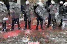 Police officers with shields, surrounded by red powder and trash on the street. sign on the ground says in Serbian: “He knows that we know that he is finished”