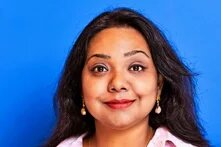 Person looks directly at the camera, long dark hair, earrings, blue background.