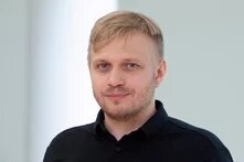Mann mit kurzem blondem Haar und Bart schaut in die Kamera, steht vor heller, unscharfer Wand.