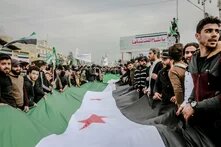 Viele Menschen halten gemeinsam eine große Flagge mit Streifen in Grün, Weiß und Schwarz sowie drei roten Sternen in der Mitte.