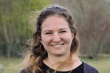 Person mit langen, teils gelockten Haaren steht draußen vor Bäumen und Wiese, schaut lächelnd in die Kamera.