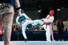 Two taekwondo fighters exchange kicks mid-air during a match; one wears blue gear, the other red. Judges and spectators in the background.