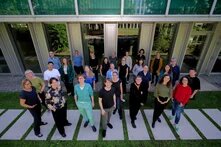 Gruppe von etwa 30 Personen steht draußen vor einem Gebäude auf Steinplatten in Gras, frontal zur Kamera blickend.