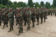 Group of soldiers in camouflage uniforms with rifles marching on sandy ground, trees in the background.