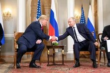 Donald Trump and Vladimir Putin sit facing each other and shake hands. A small table with flowers is between them.