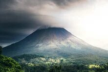 A lone volcano rises from dense vegetation, its peak covered by dark clouds.