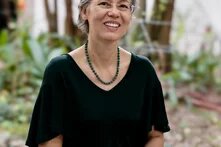 Person with short wavy gray hair, glasses, and green necklace sits outdoors smiling, hands resting on patterned trousers.