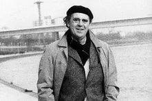 Heinrich Böll mit Mantel und Mütze geht lächelnd am Fluss Rhein entlang; im Hintergrund eine Brücke und Industrieanlagen.