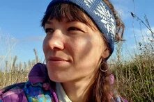Person looks to the left, wears a patterned headband, and stands outdoors with dry vegetation and blue sky in the background.