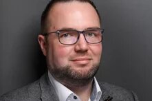 Person mit Brille vor einfarbigem Hintergrund, leicht zur Seite gedreht und mit neutralem Gesichtsausdruck.