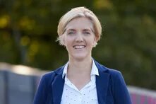 Person mit kurzem, blondem Haar steht draußen und blickt lächelnd in die Kamera. Unscharfer Hintergrund mit Bäumen.